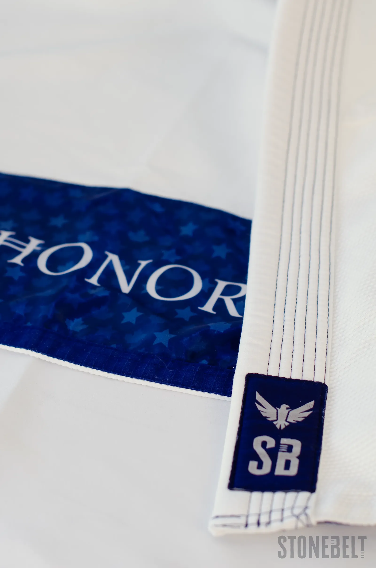 Close-up of Stonebelt Heritage BJJ gi showing navy SB eagle woven patch on lapel, multi-row reinforced stitching, and navy maple leaf interior lining with Honor branding