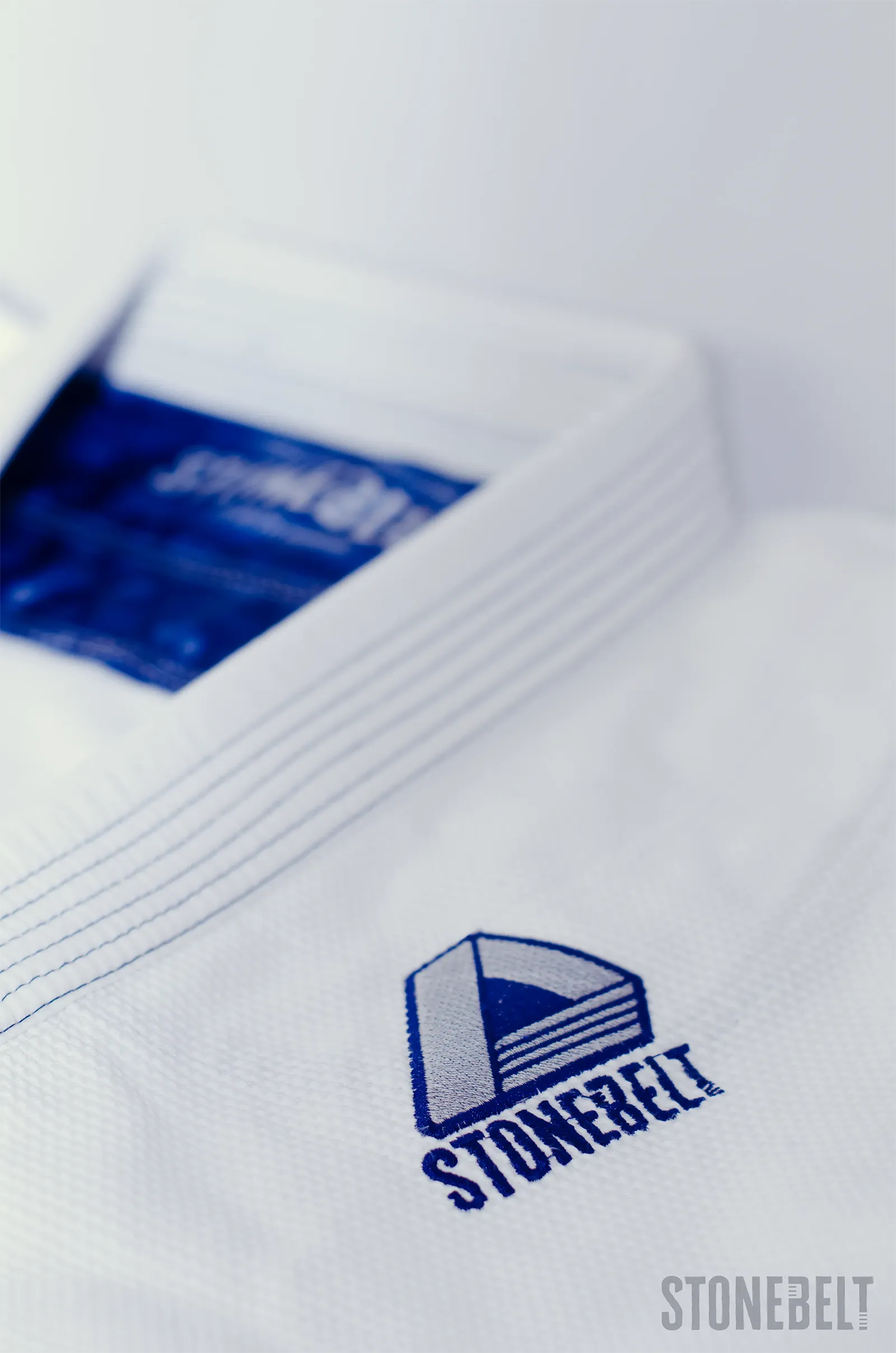 Macro close-up of Stonebelt Heritage BJJ gi showing navy embroidered diamond chest logo with Stonebelt wordmark, reinforced multi-row lapel stitching, and interior navy branded label