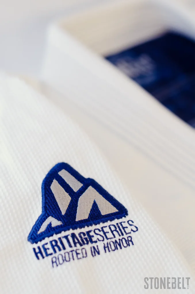 Macro close-up of Stonebelt Heritage BJJ gi sleeve showing embroidered Heritage Series Rooted In Honor shoulder patch with mountain logo on pearl weave fabric