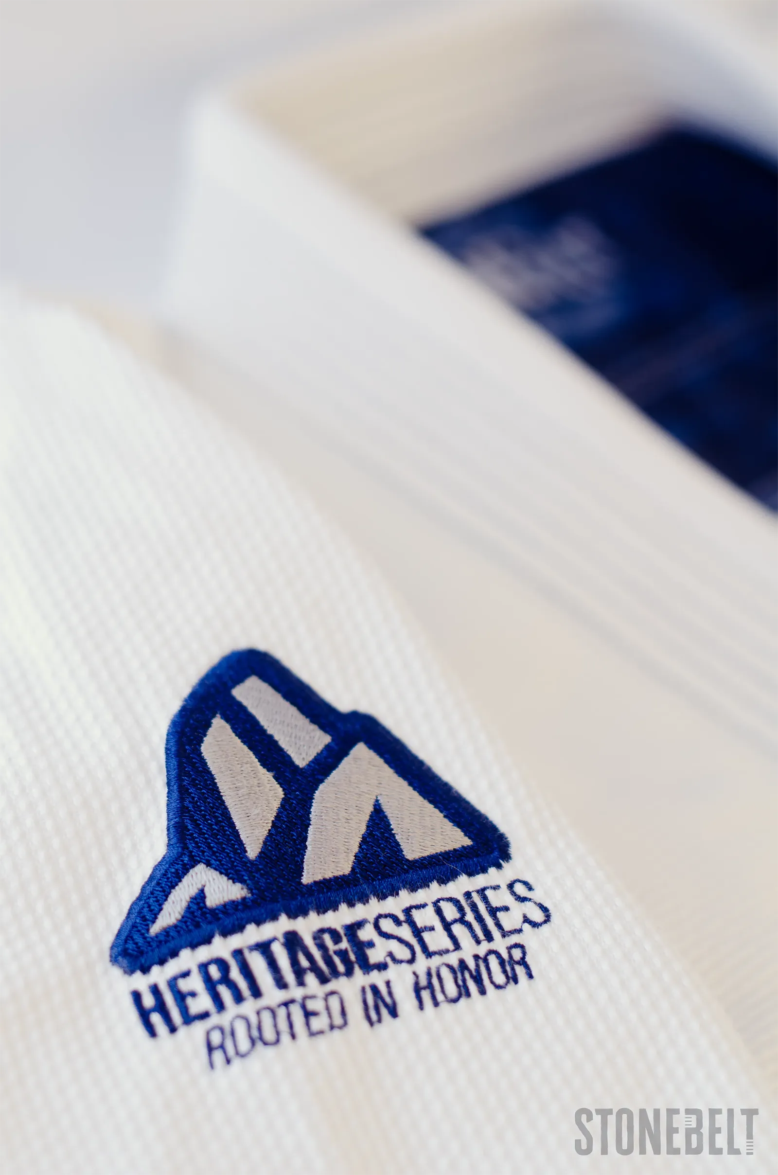 Macro close-up of Stonebelt Heritage BJJ gi sleeve showing embroidered Heritage Series Rooted In Honor shoulder patch with mountain logo on pearl weave fabric