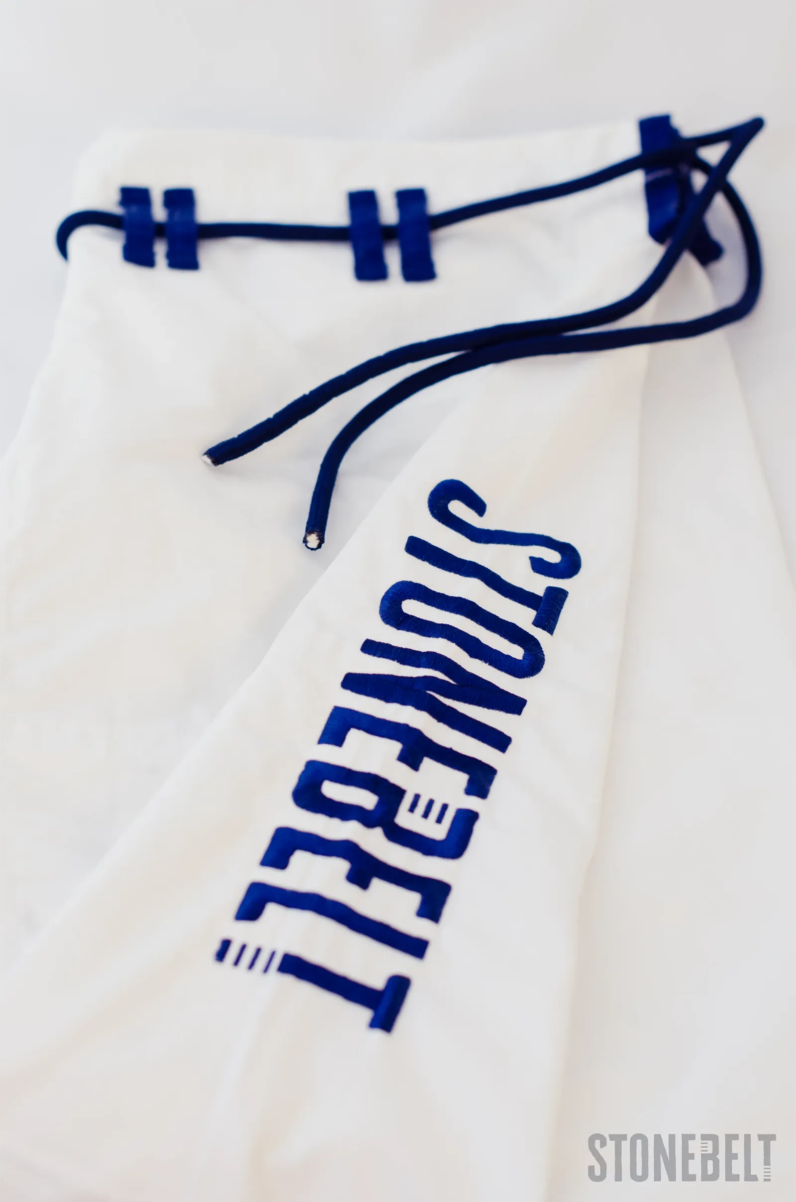 Close-up of Stonebelt Heritage BJJ gi 10 oz ripstop pants showing navy blue embroidered Stonebelt logo, reinforced drawstring loops, and navy drawstring cord