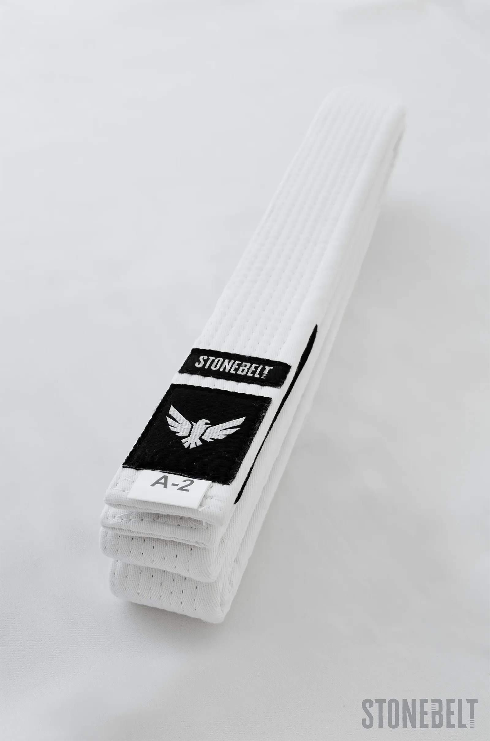 Close-up of folded Stonebelt BJJ white belt showing eagle logo patch, rank bar for stripe progression, and A-2 size label