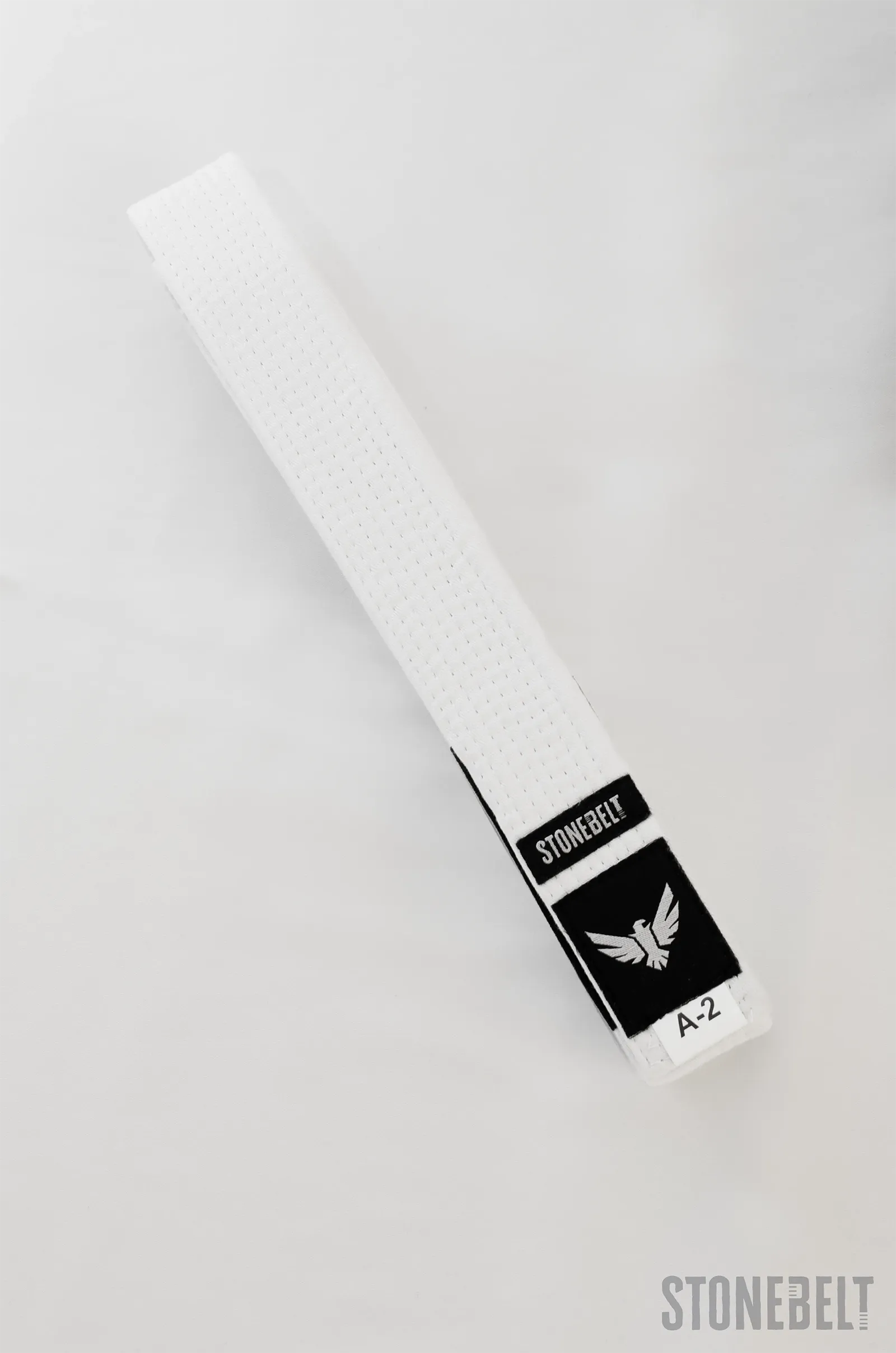 Stonebelt BJJ white belt cotton rank belt laid flat showing full length, Stonebelt eagle logo patch, traditional rank bar, and A-2 size tag
