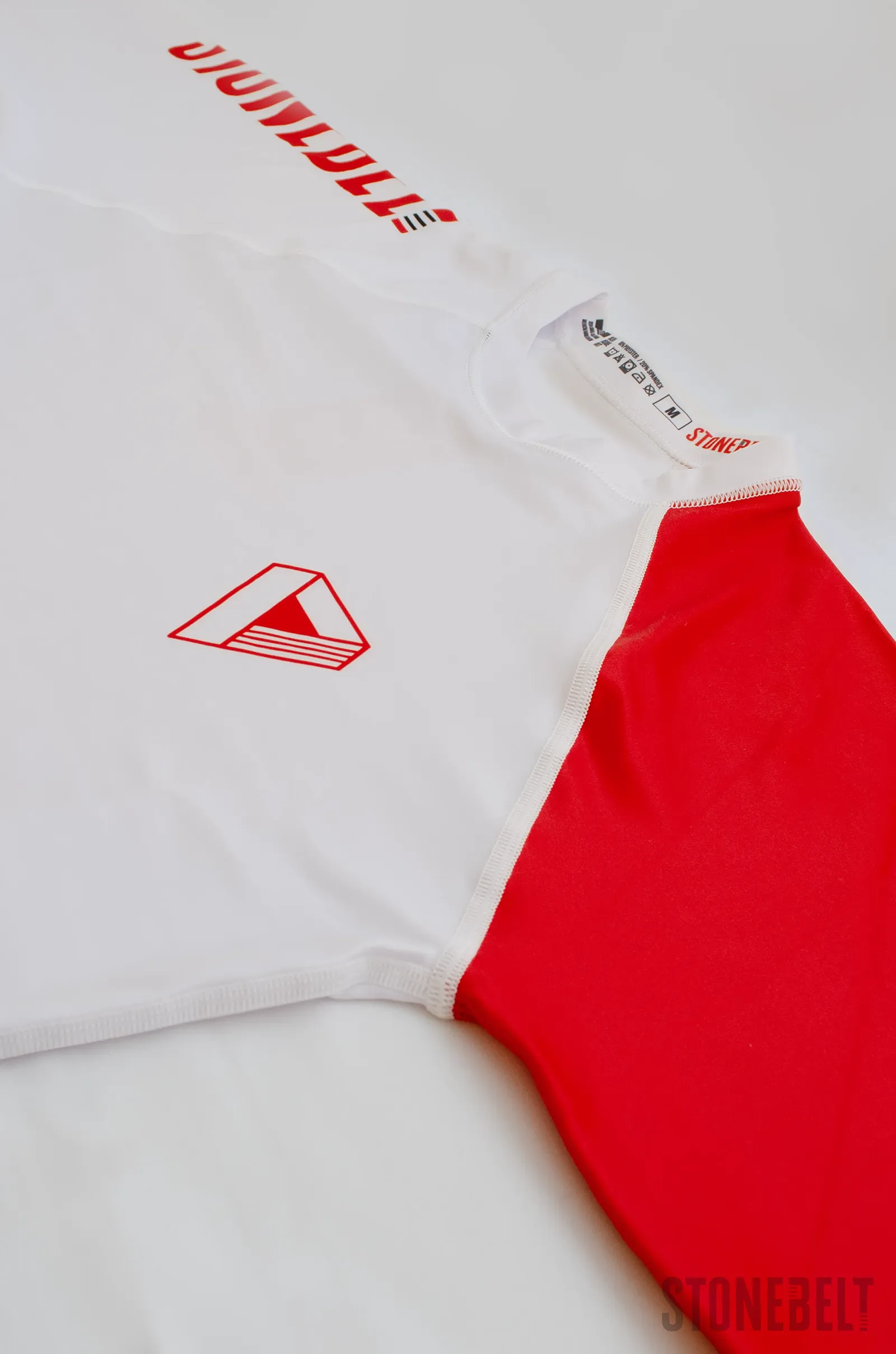 Close-up of Stonebelt Heritage long sleeve rashguard showing flatlock stitching at shoulder seam, red diamond chest logo, branded neck label with size and care instructions, and white-to-red sleeve panel transition