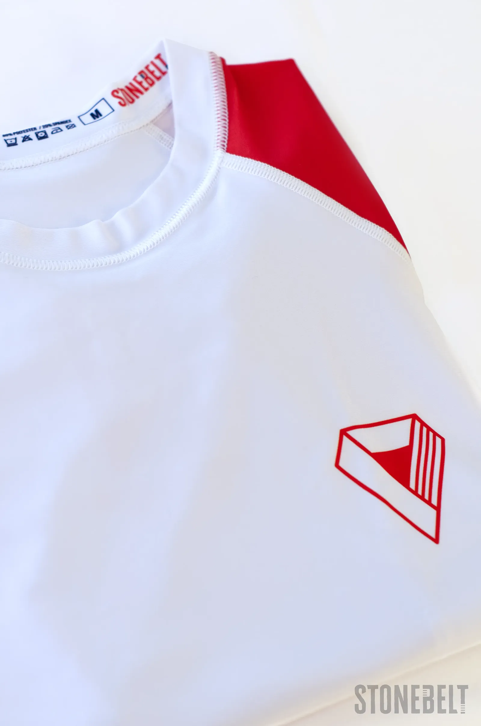 Flat lay detail of Stonebelt Heritage rashguard showing chest logo, Stonebelt branded collar tag, and red raglan shoulder