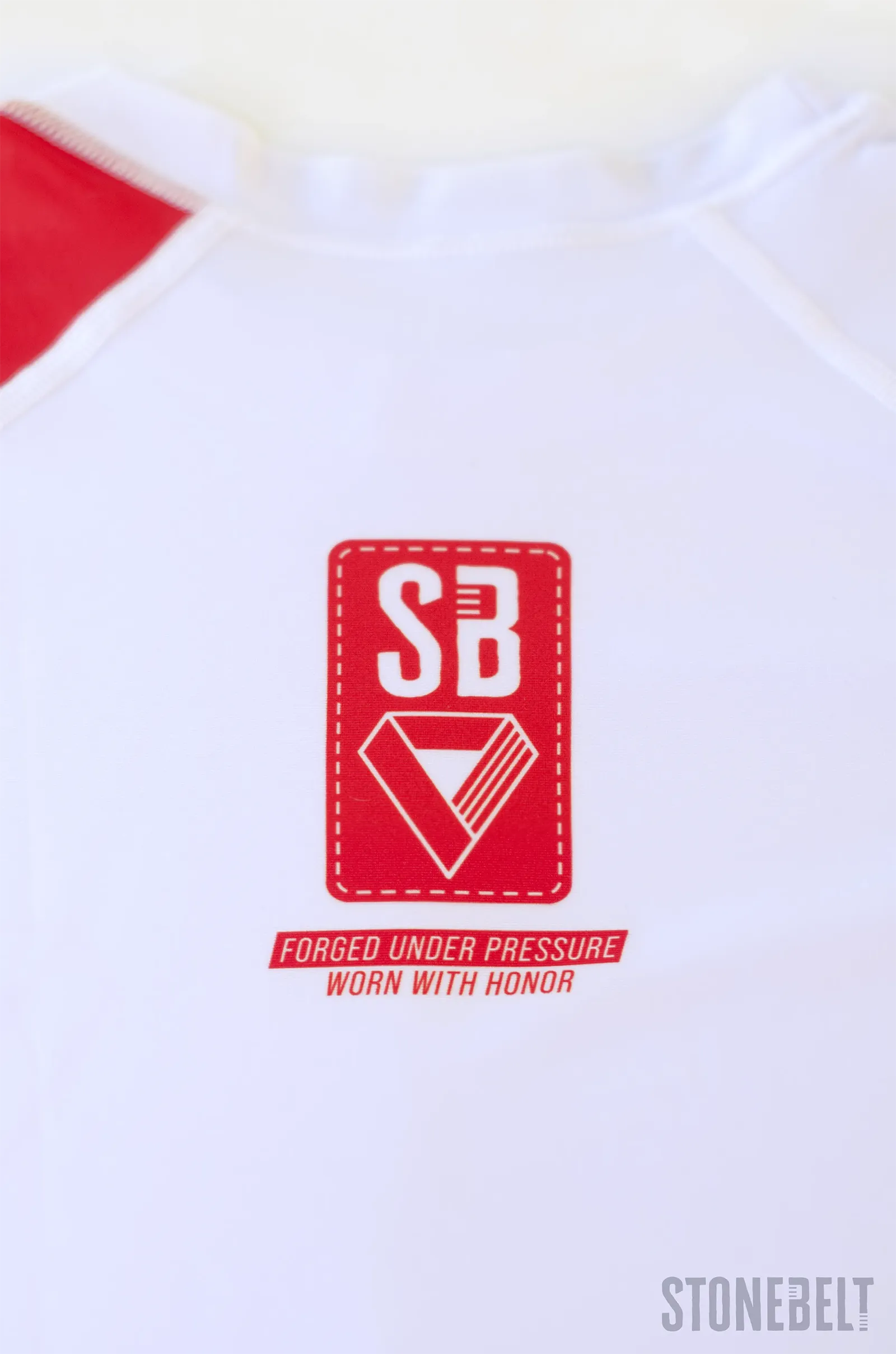 Close-up of Stonebelt Heritage rashguard front showing SB logo badge and Forged Under Pressure text on white fabric