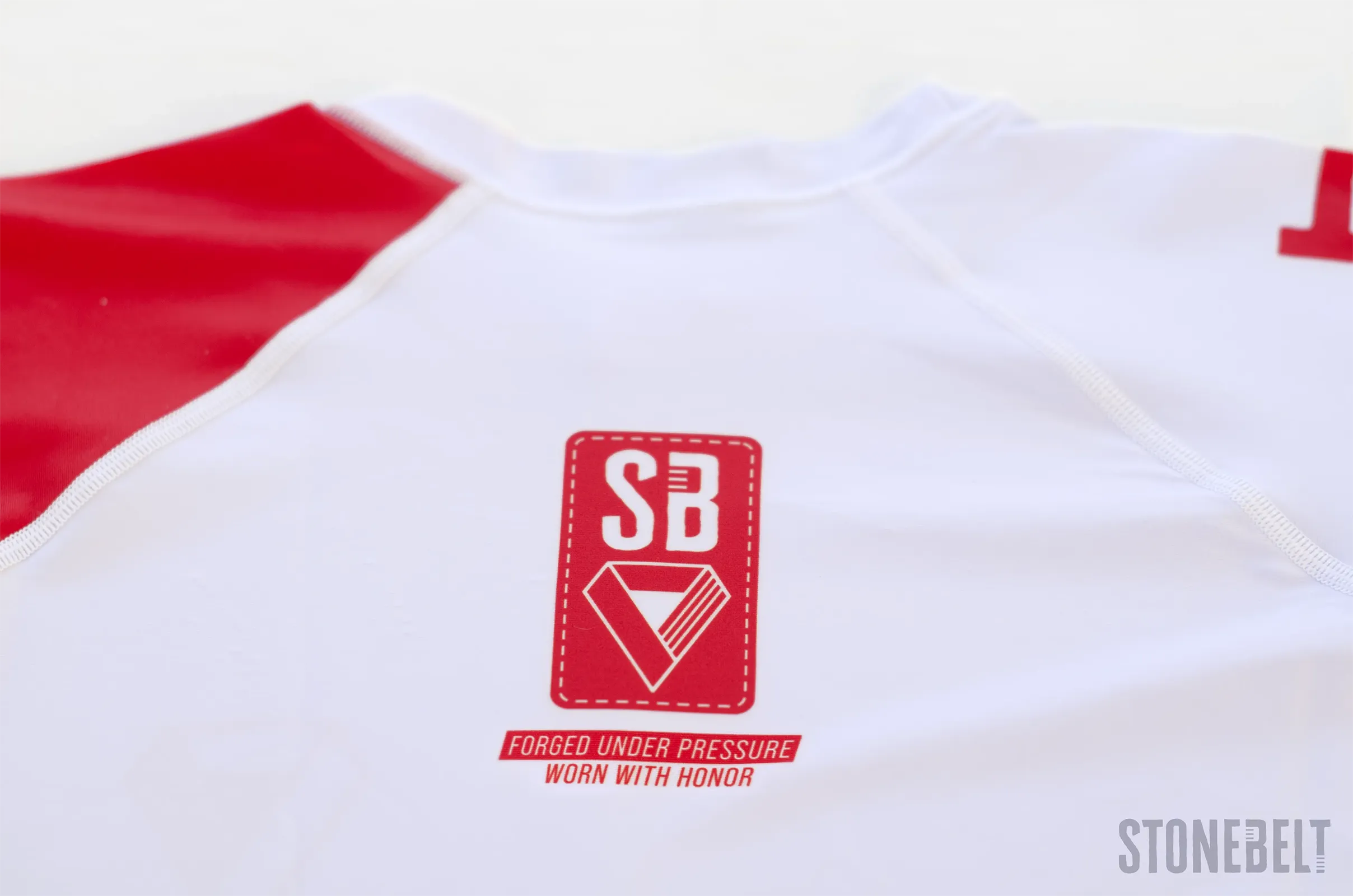 Stonebelt Heritage rashguard flat lay showing SB badge, red raglan shoulder panel, and flatlock stitching