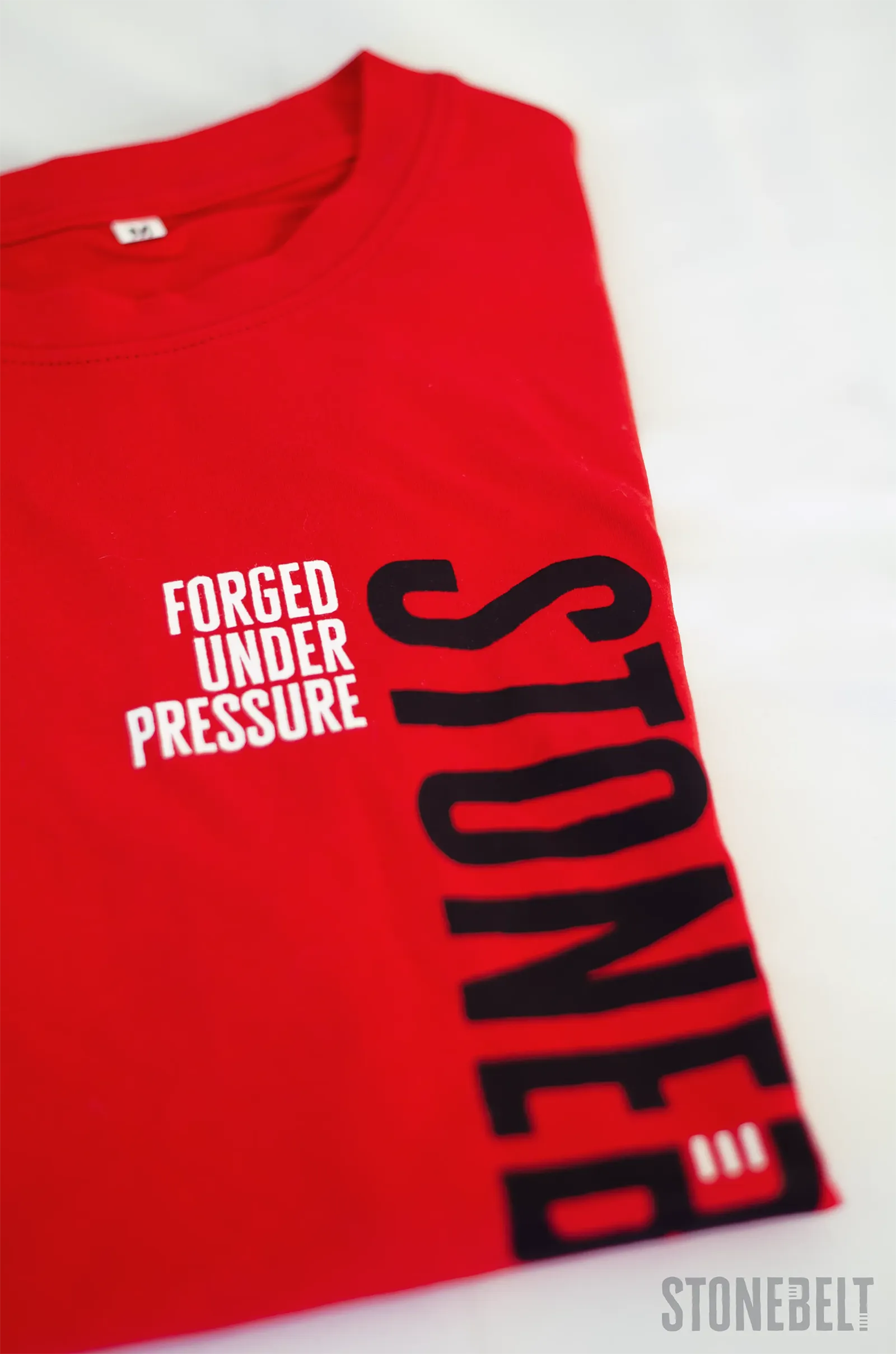 Flat lay of Stonebelt Classic Logo Tee in red showing Forged Under Pressure text and vertical Stonebelt branding