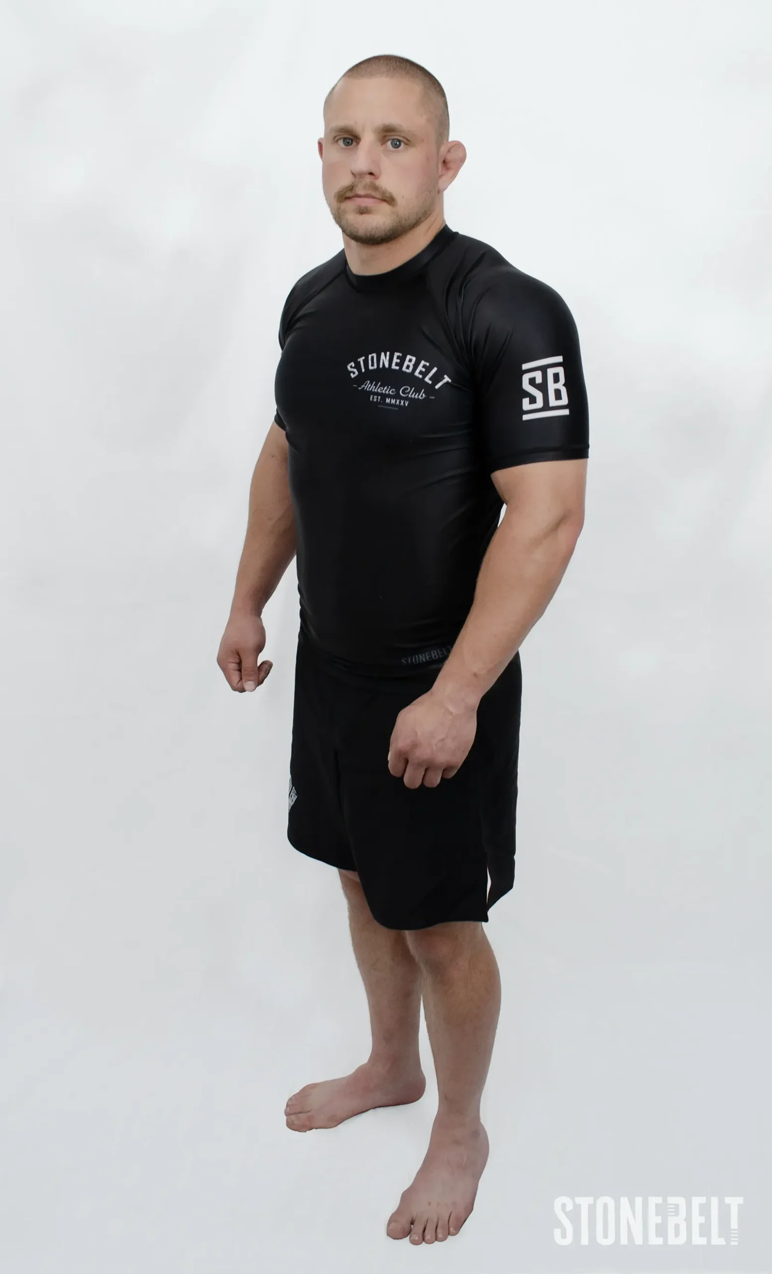 Model in Stonebelt Athletic Club black rashguard and black Heritage grappling shorts, right side angled view with SB sleeve logo
