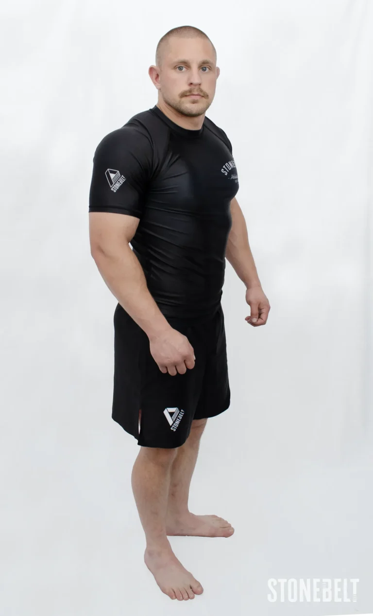 Model in Stonebelt Athletic Club black rashguard and black Heritage grappling shorts, left side angled view with shoulder logo