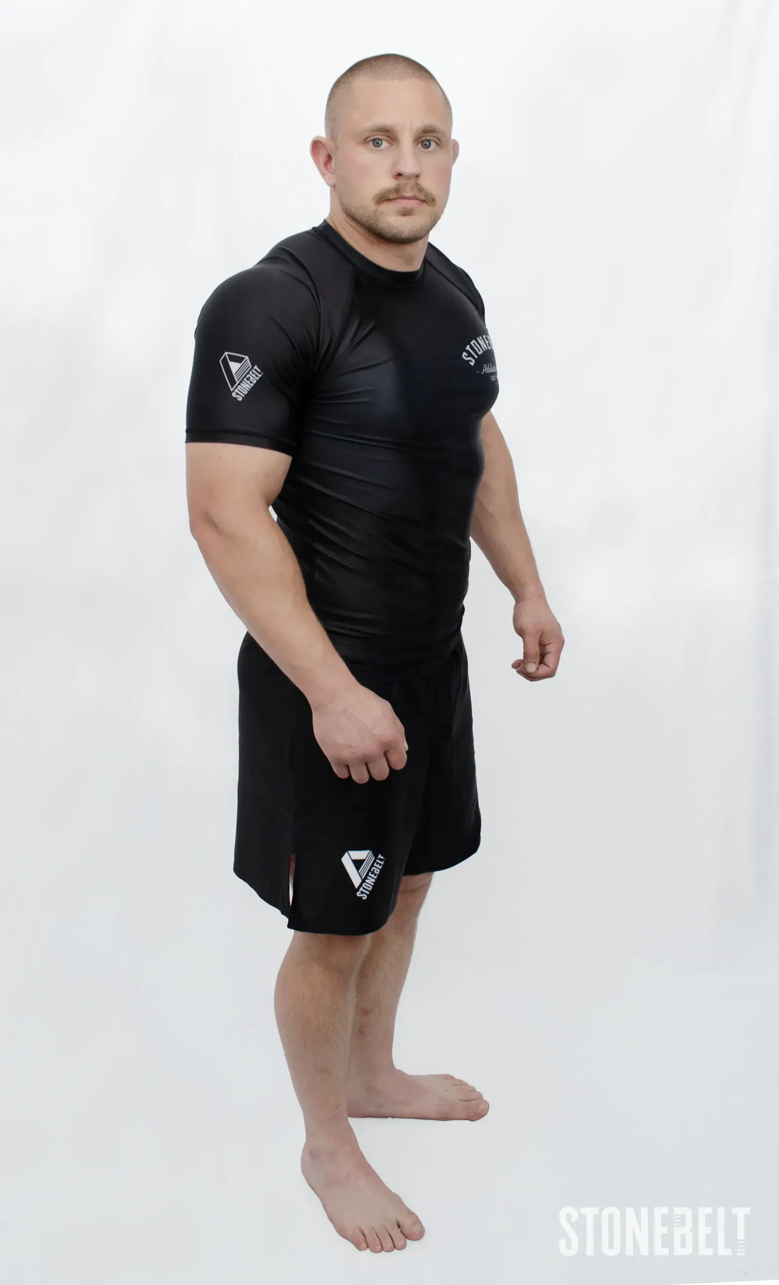 Model in Stonebelt Athletic Club black rashguard and black Heritage grappling shorts, left side angled view with shoulder logo